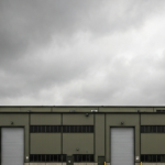 Rüstungsfabrik-Außengelände unter bedecktem Himmel in gedeckten Militärgrün- und Grautönen