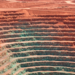 Luftaufnahme eines Kupfer-Tagebaus mit rötlich-orangefarbenen Felsterrassen und blaugrünen Oxidationszonen in Nordchile