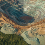 Luftaufnahme eines Kupfertagebaus mit rötlich-orangen Terrassenstufen und grün oxidiertem Gestein in hügeliger Landschaf
